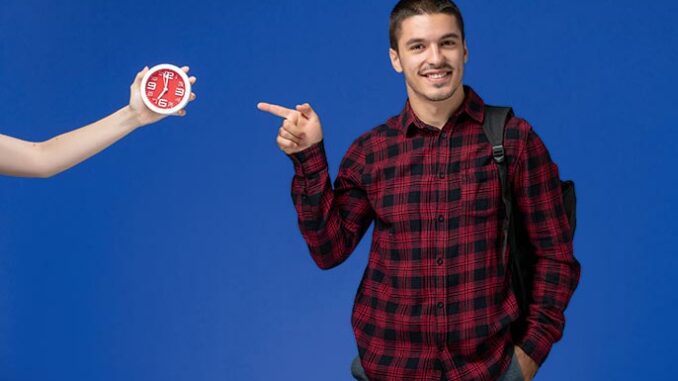 vorderansicht-des-maennlichen-studenten-im-roten-karierten-hemd-mit-dem-rucksack-der-auf-der-blauen-wand-laechelt